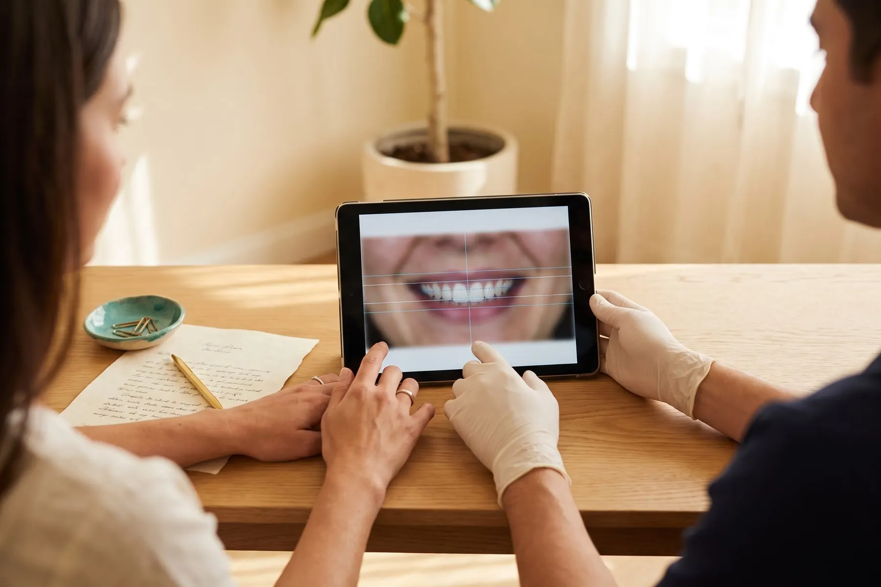 Las manos de un doctor y de una paciente sobre una tablet con una vista previa del diseño de sonrisa, junto a un presupuesto impreso y un bolígrafo dorado